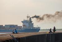 Smoke pours from a cargo ship sailing past a group of fishermen on a pier.