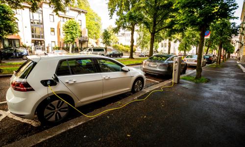 An electric car is charging on the side of a tree-lined road.