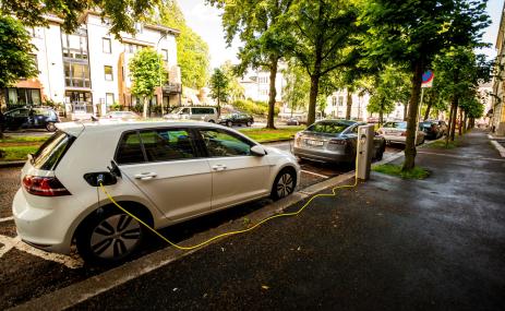 An electric car is charging on the side of a tree-lined road.