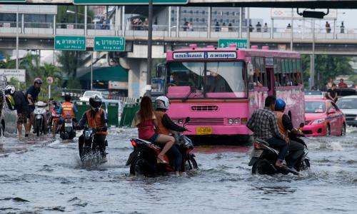 Motorcyclists, buses and pedestrians navigate a flooded, hectic city street.