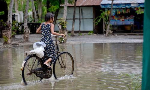 A girl wearing a long sundress pedals a bicycle down a flooded road..