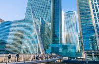 People walk across a bridge toward tall glass bank buildings.