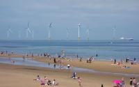 Groups of people are scattered across a large, sandy beach on a sunny day with floating wind turbines in the background.