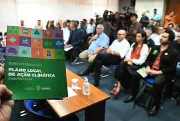 A pamphlet about Campinas's climate action plan is held up before a city hall audience.