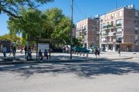 People wait at a bus top with trees and apartment buildings in the background.