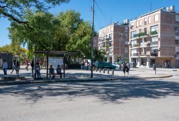 People wait at a bus top with trees and apartment buildings in the background.