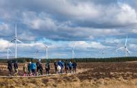 People walking on a boardwalk with trees and wind turbines in front of them.